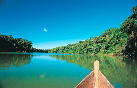 A wooden canoe pointing towards a calm river surrounded by lush green forest under a clear blue sky.