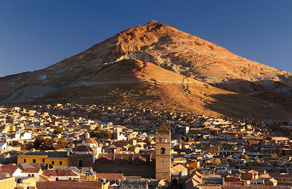 A sunny panorama of a town at a mountain's base, featuring densely packed buildings and a prominent church tower.