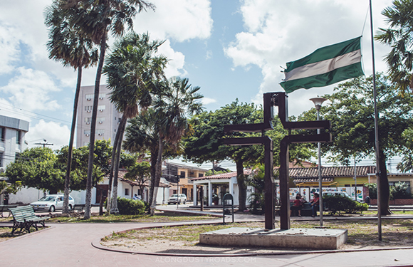 A public square image featuring a large cross, palm trees, a flagpole, buildings, under a partly cloudy sky.