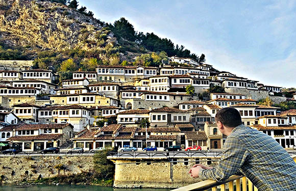 A man in checkered jacket observes a hillside of traditional white houses in a historical town by water.