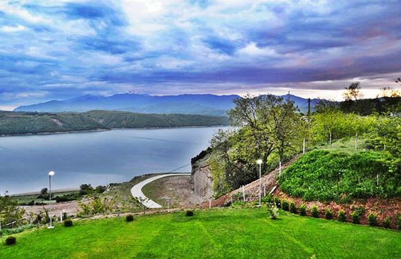 A lush lawn and terraced hillside overlook a calm lake, with distant mountains under a dramatic, colorful sky.