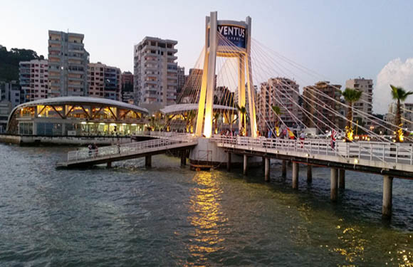 A pedestrian bridge with a modern design featuring cables and a tower, illuminated at dusk, with city buildings in the background.