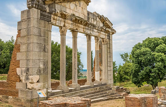 A photo of ancient ruins with several standing columns and an architrave, set against a cloudy sky with greenery in the background.