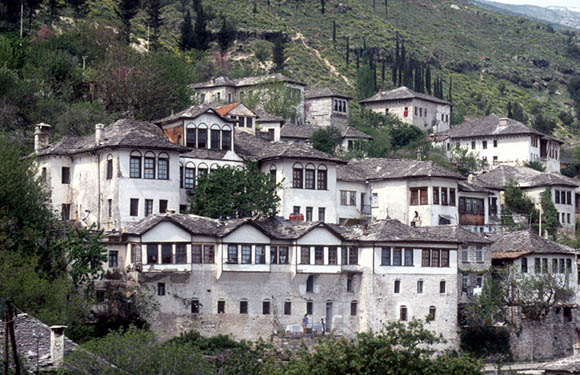 A cluster of traditional white houses with dark roofs on a hillside with trees in the background.