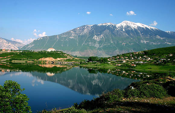 A scenic view of a calm lake reflecting the image of a village and snow-capped mountains under a blue sky with scattered clouds.