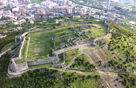 Aerial view of a large, ancient fortress with ruined walls and structures spread over a green landscape with a city in the background.