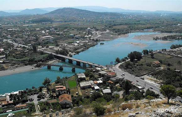 Aerial view of a landscape featuring a river with bridges, surrounded by buildings and greenery with mountains in the background.