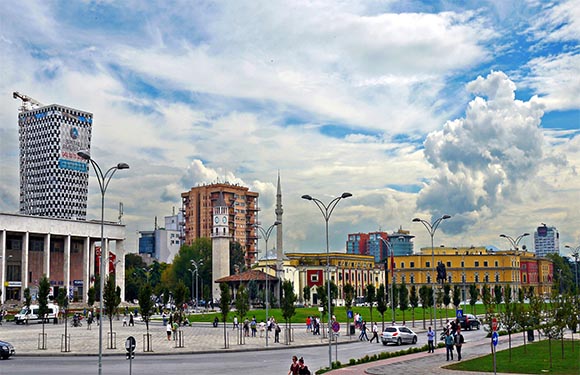 A cityscape with modern buildings under a partly cloudy sky, a busy street in the foreground, and pedestrians crossing the road.