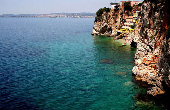 A coastal landscape with clear turquoise waters beside a rocky cliff with buildings perched on the edge.