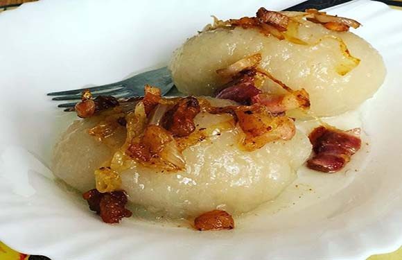 A plate with two potato dumplings topped with crispy bacon bits and caramelized onions, with a fork resting on the side.