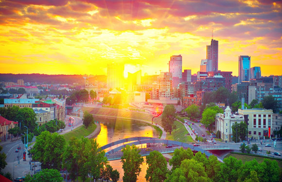 A vivid sunset silhouettes city buildings, with a river in the foreground and trees dotting the urban scene.