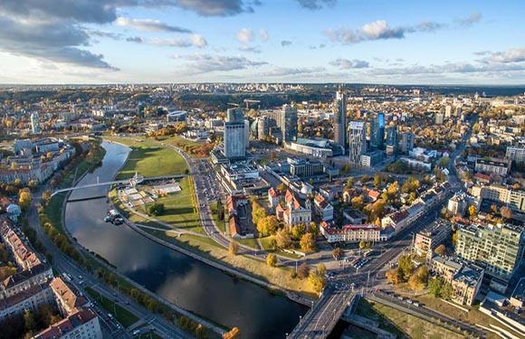 Aerial view of a city with skyscrapers, a central river, green spaces, and mixed-use buildings under a partly cloudy sky.