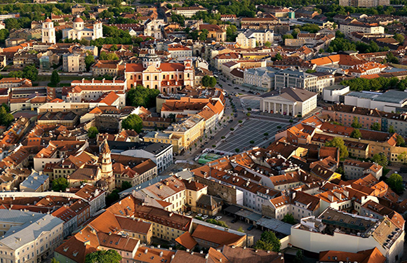 Aerial view of a densely populated European city with historic architecture, red-roofed buildings, streets, and a large square.