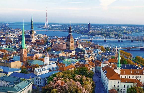 Aerial view of a city with historic architecture, multiple spires, a river, bridges, and green spaces.