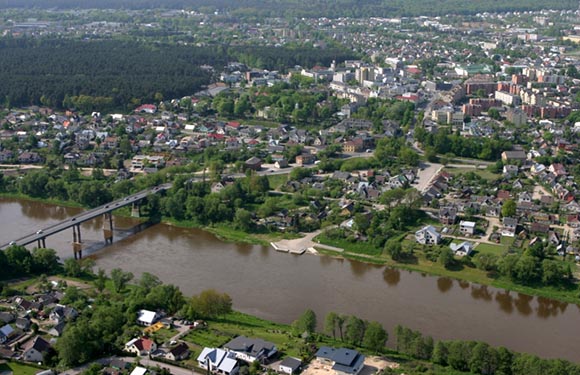 Aerial view of a town with dense housing and greenery, intersected by a river with a bridge crossing over it.