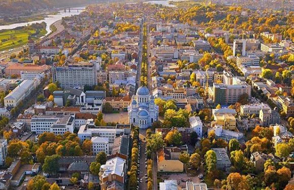 Aerial view of a city with dense buildings, a domed structure, tree-lined streets, and a river, bathed in warm, golden light.