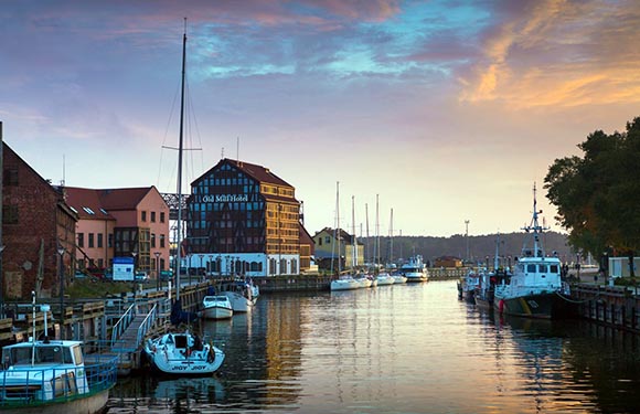 A scenic view of a tranquil harbor at sunset with boats docked along the water, adjacent to buildings with a colorful sky in the background.