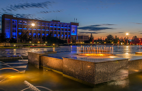 An image of a lit fountain at dusk with a building in the background and lights reflecting on the water.