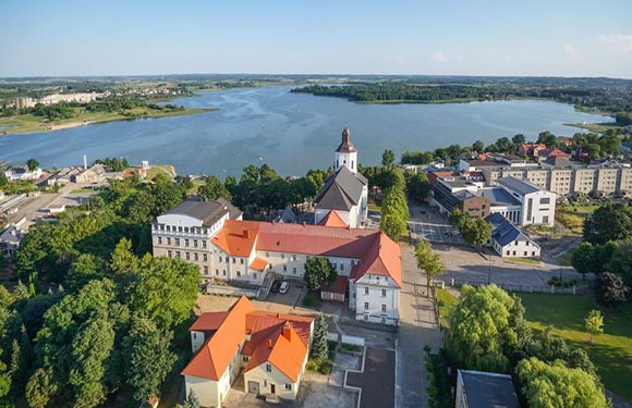 Aerial view of a town with historic buildings near a large river, greenery surrounding the area, under a clear sky.