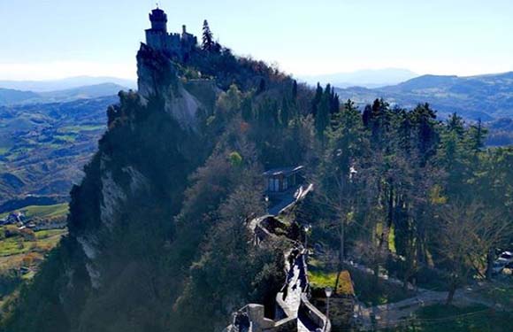 A castle perched on the peak of a steep hill with surrounding greenery and a clear sky.