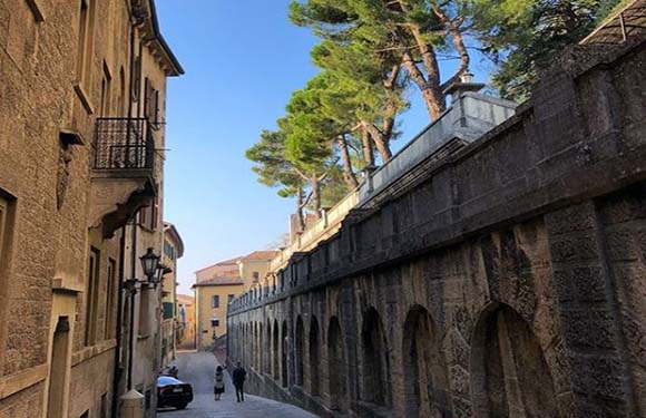 A narrow street flanked by old buildings and a stone aqueduct, with visible trees, pedestrians, and a distant parked car.