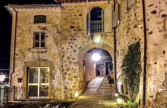 A night-time view of an old stone building with an arched passageway leading to a staircase, illuminated by warm lights.