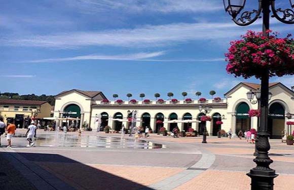 A public square scene with people, a building with an arched entrance, and a flower-adorned street lamp.