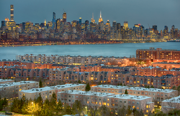 A cityscape at dusk with illuminated buildings and a skyline in the background, foreground showing residential buildings with lights on.