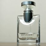 A clear glass perfume bottle with a silver cap on a beige background, labeled "BVLGARI" with additional text that is not clearly legible.