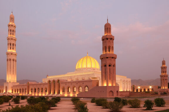 A mosque with two tall minarets and a large golden dome, illuminated at twilight, with a clear sky in the background.