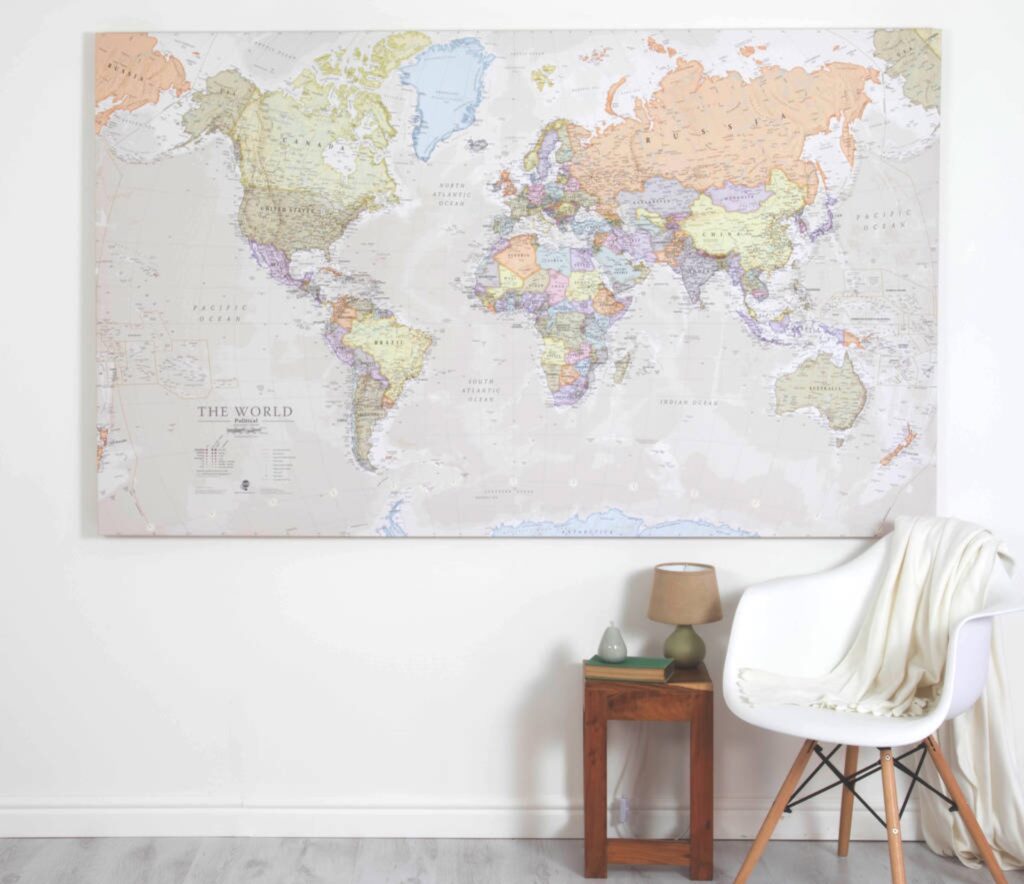 A world map canvas hangs above a table with a vase and books, beside a white chair with a blanket.