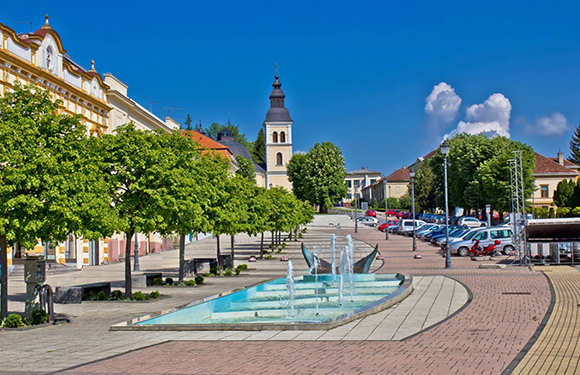 A sunny day in a town square with a fountain, parked cars, trees lining the streets, and a church tower in the background.