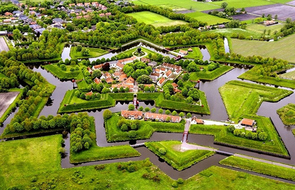 Aerial view of a star-shaped fort with grassy walls, red-roofed buildings, water moats, and countryside backdrop.