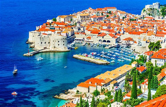 Aerial shot of a coastal town with historic buildings and a fortress, surrounded by the sea with docked boats.