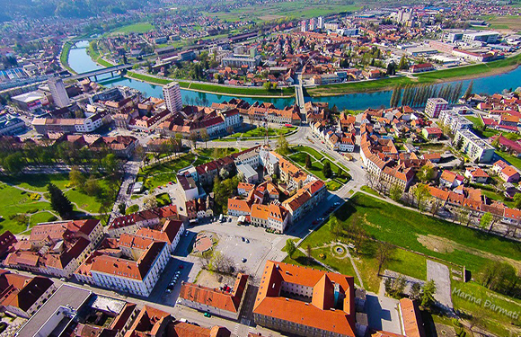 Aerial view of a historic town with red-roofed buildings, streets, green spaces, and a river running through it.