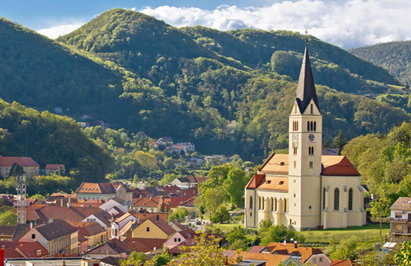 A picturesque view of a small town nestled in a valley with a prominent church spire, surrounded by lush green hills under a partly cloudy sky.