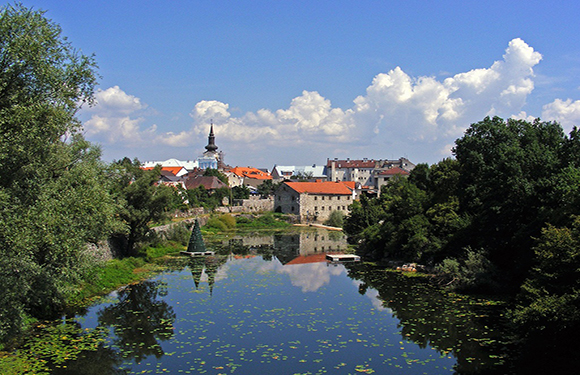 A scenic view of a calm river with reflections of trees and buildings, a town with historic architecture under a blue sky with fluffy white clouds.