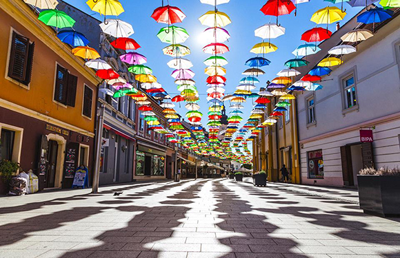 A vibrant array of umbrellas hangs above a pedestrian street, casting shadows under a clear blue sky.