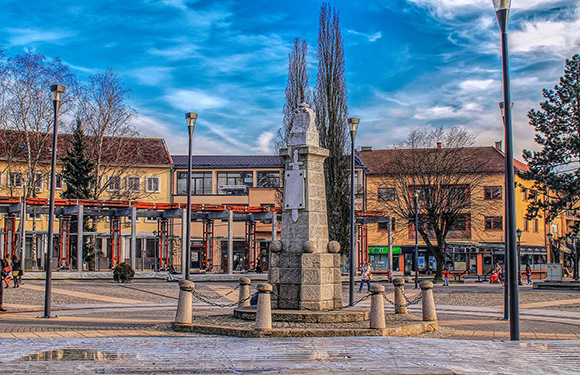 A town square features a central monument, benches, cobblestone ground, buildings, bare trees, under a partly cloudy sky.