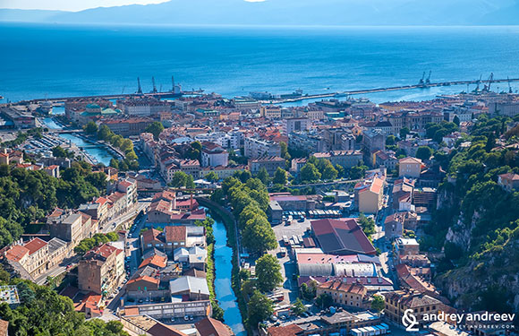 Aerial view of a coastal city with dense buildings and streets, with the sea in the background and hills in the foreground.