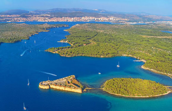 Aerial view of a coastal landscape with clear blue waters, several boats, a green forested peninsula, and a city in the distance.