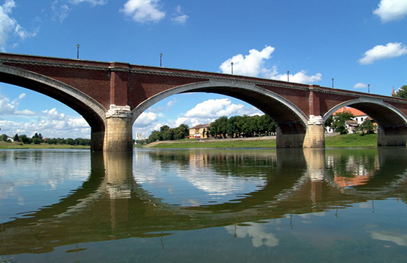 A multi-arched bridge crosses a calm river, under clear skies, with buildings in the background.