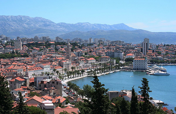 A coastal city with red-roofed buildings, a promenade, docked boats, mountains, and clear sky.