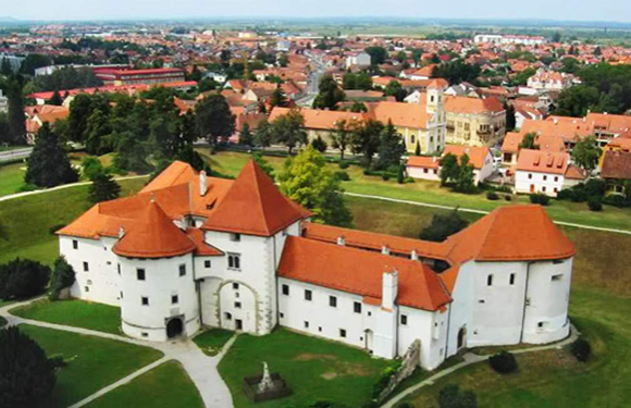 Aerial view of a large white castle with red roofs surrounded by green lawns and trees, with a town in the background.