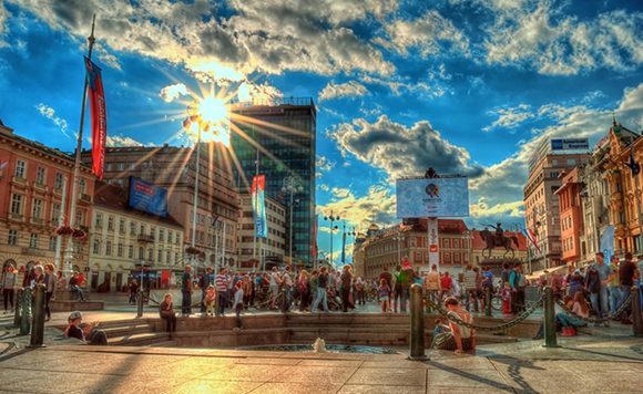 A lively city square filled with people, buildings, a clear blue sky with clouds, and bright sunshine.