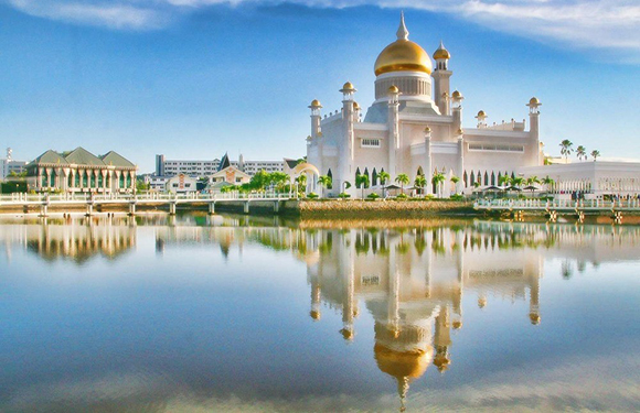 A mosque with a large golden dome and several smaller domes, reflected in a body of water under a clear sky.