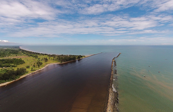 Aerial view of a thin land strip separating a brown river and blue sea, with visible vegetation and a partly cloudy sky.