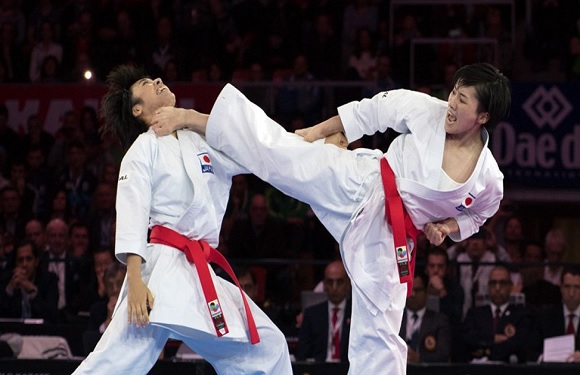 Two people in white karate uniforms and red belts are in a martial arts demo, with one performing a high kick.