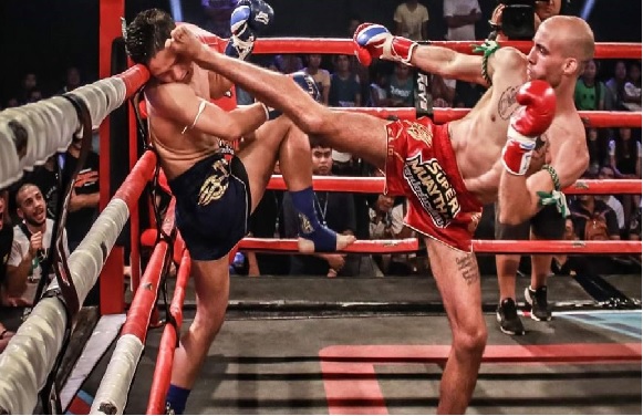 Two Muay Thai fighters in a ring, one landing a high kick on the other's head. Spectators are visible in the background.