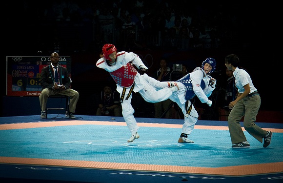 Two taekwondo athletes spar; the red-gear athlete kicks at the blue-gear one, under a referee's watch on a blue mat.